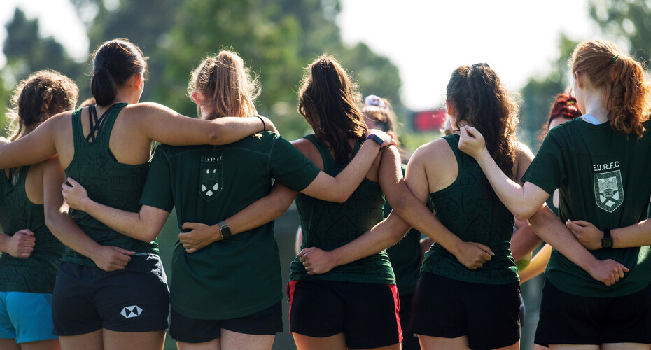 Seen from behind, a female rugby team huddle with their arms round each others' waists