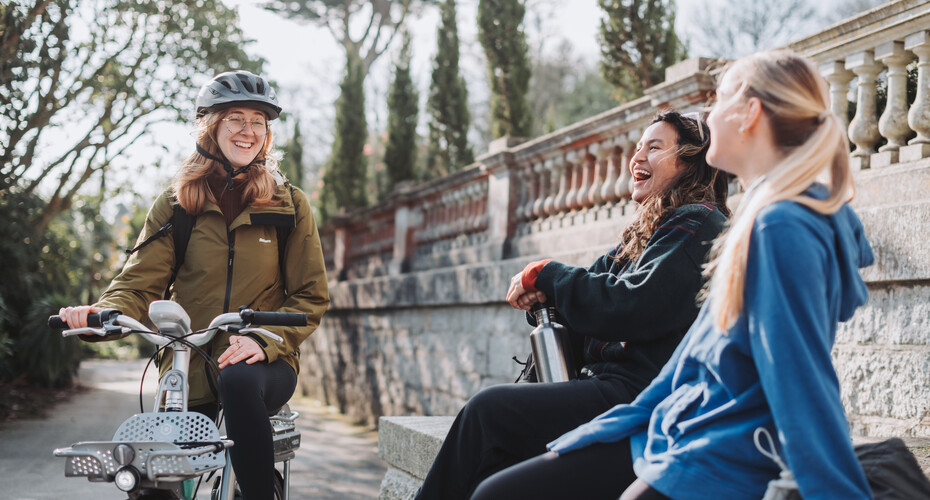 Three students laughing together in Reed Hall grounds. One has a bicycle with her and is wearing a helmet.